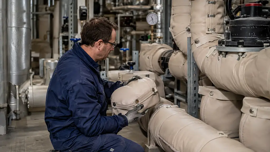 maintenance technician inspecting a removable insulated valve cover beside precisely fitted insulation around pipe elbows in a narrow industrial plant room, editorial photography, no text, no logo