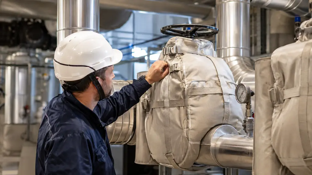 Maintenance technician inspecting removable insulation jackets on steam valves and metal-clad insulated pipes inside a clean industrial boiler room, realistic editorial photography, no text, no logo