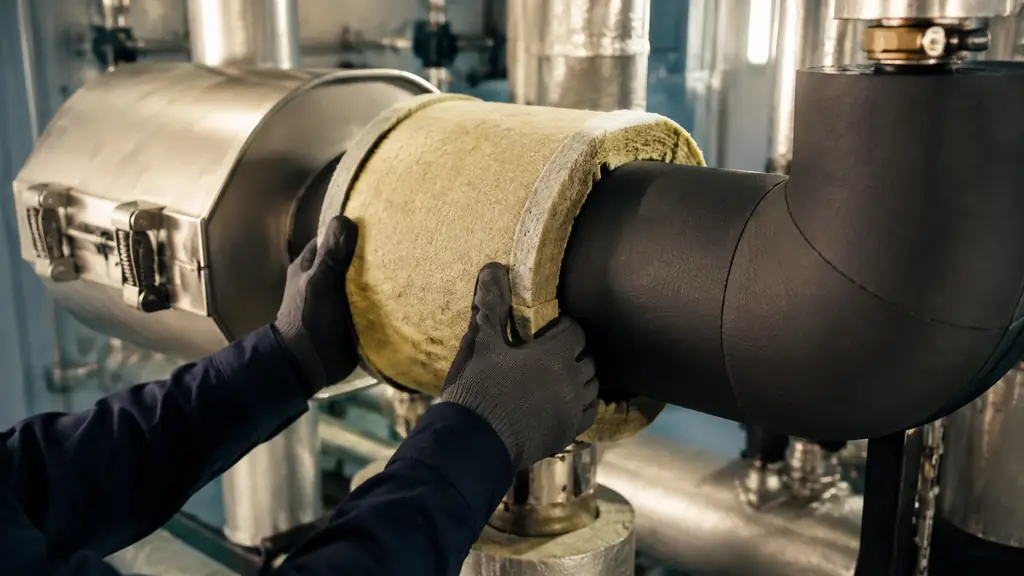 Close-up editorial photograph of mineral wool pipe insulation being fitted beside a black elastomeric foam elbow, with a removable metal-clad valve cover in an industrial boiler room, no text, no logo