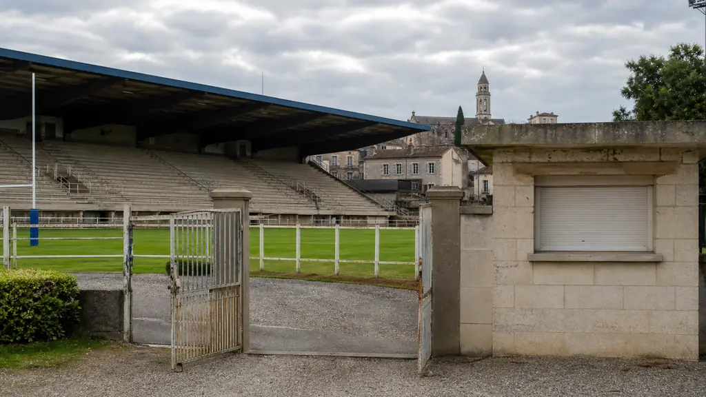 Editorial photograph of an empty rugby stadium in Périgueux, vacant green pitch, closed ticket booth and unoccupied stands, overcast summer afternoon, no text, no logos