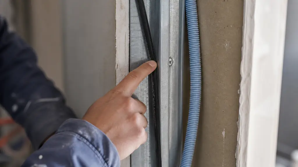 Close-up editorial photograph of an electrician inspecting an unprotected R2V cable pinched against a metal stud inside an open plasterboard wall, with an ICTA conduit and mounting screw visible nearby, no text, no logo