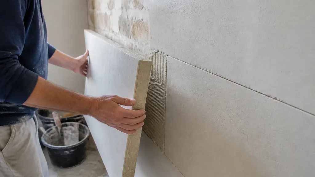 Close-up editorial photograph of a calcium silicate board measuring 1000 by 610 by 50 mm being fitted onto a cold interior masonry wall, with visible tight joints and mineral adhesive, no text or logo