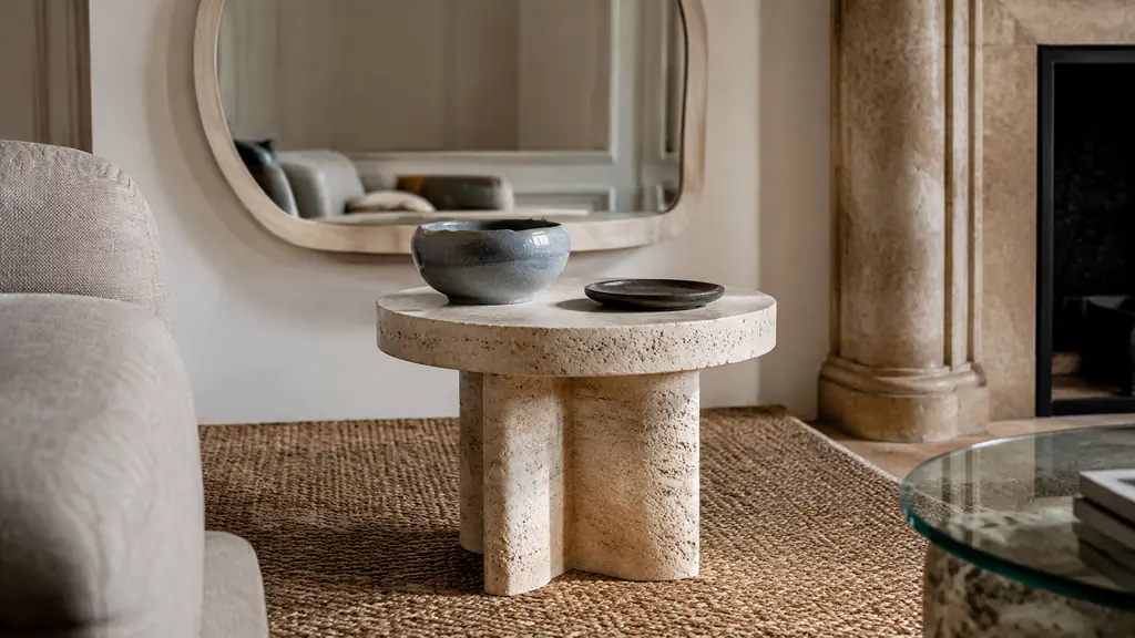 editorial close-up of a living room corner with a travertine side table, a woven jute rug and an organic rounded mirror, soft natural daylight, no text, no logo