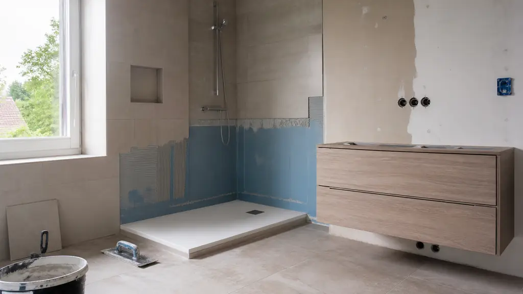 A bathroom renovation in progress showing a walk-in shower with an extra-flat tray, waterproofing beneath partially installed tiles, and a wall-mounted vanity awaiting final installation, editorial photography, no text, no logo