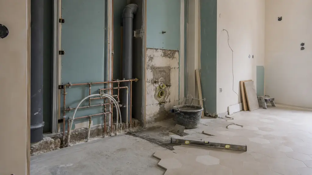 Editorial photograph of a bathroom renovation in progress, showing newly installed plumbing pipes, a VMC duct and partially laid floor tiles, no text or logo