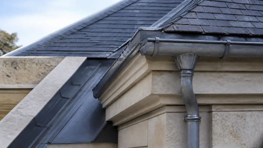 Close-up editorial photograph of a zinc box gutter inset between two roof slopes beside a stone cornice, with a hook-mounted hanging gutter and downpipe connection visible, no text, no logo